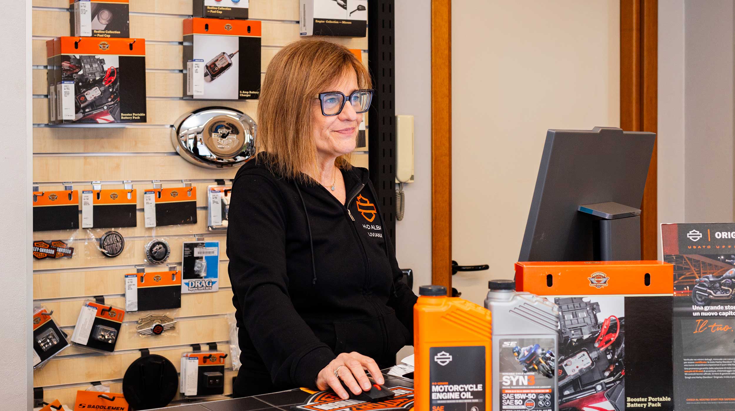 Addetta concessionaria Harley-Davidson Alba al banco ricambi e accessori, sorridente, con oli motore e prodotti originali in esposizione.