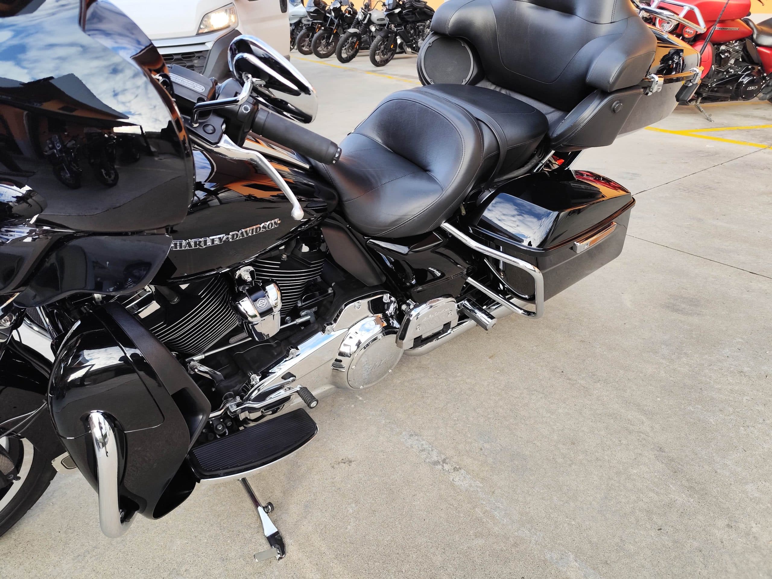 Motocicletta Harley-Davidson nera lucida parcheggiata su asfalto, con dettagli cromati, motore bicilindrico a V, sella in pelle e borse laterali. Riflessi di cielo e altri veicoli sulla carrozzeria.