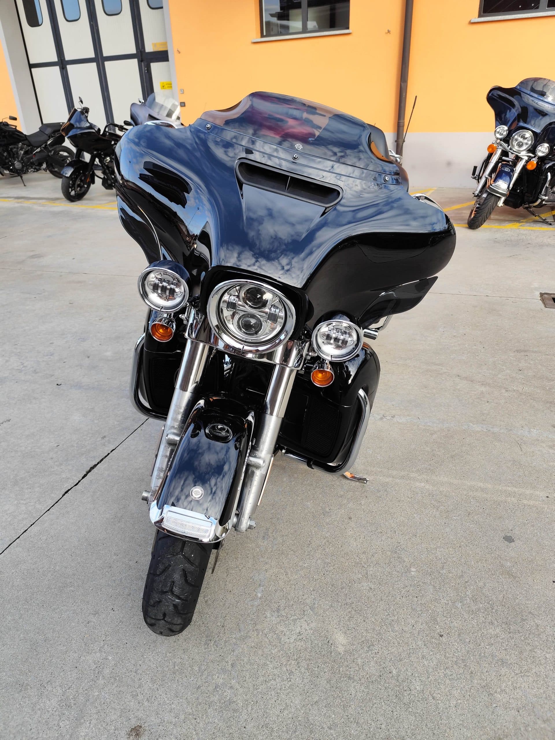 Harley-Davidson touring nera, vista frontale. Carenatura lucida riflette cielo e nuvole. Fari LED, cromature. Altre moto per test ride e concessionaria.
