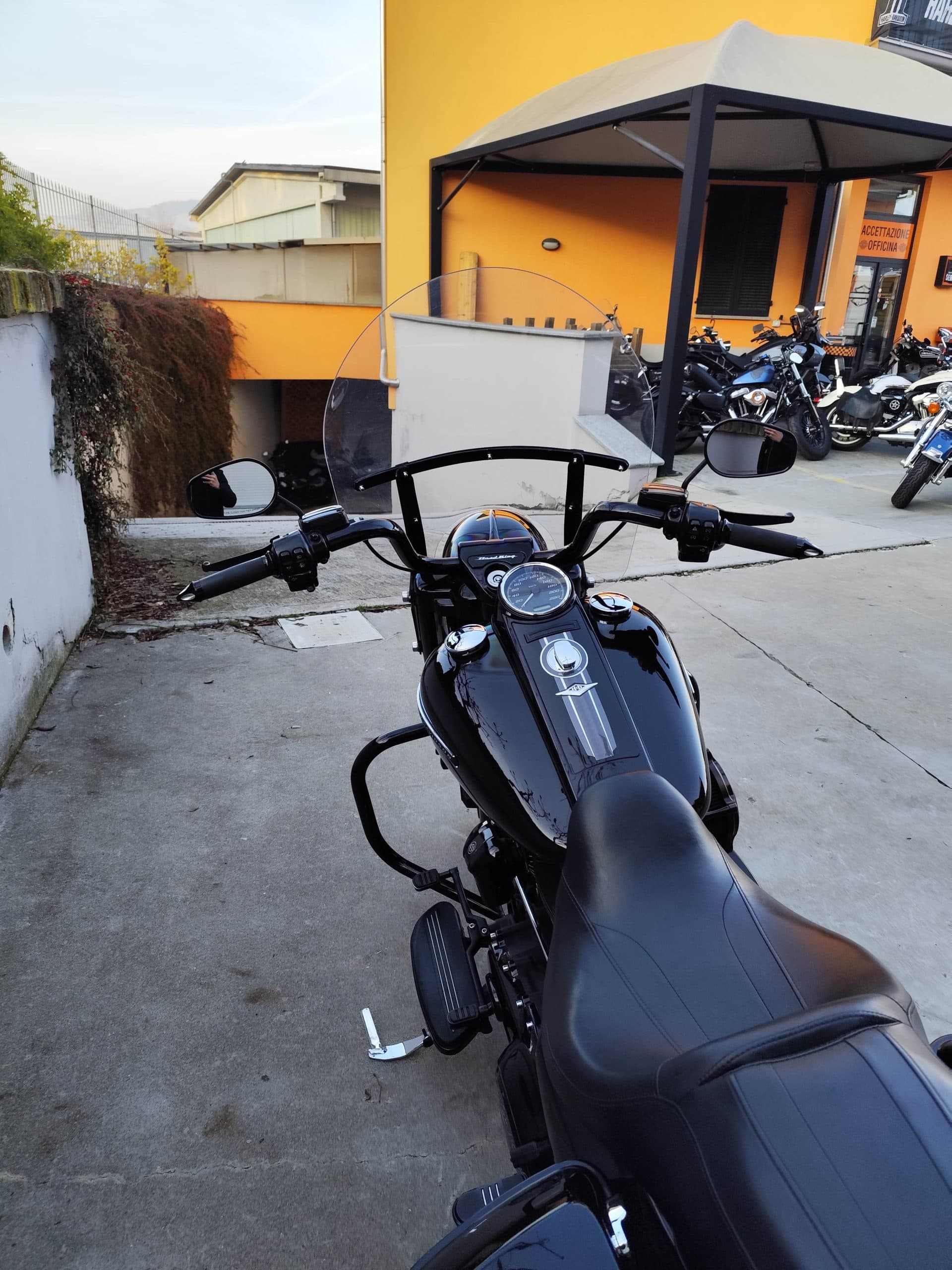 Harley-Davidson nera, vista dal posto di guida, con manubrio, parabrezza e serbatoio lucido. Sfondo: piazzale di concessionaria con officina e altre moto.