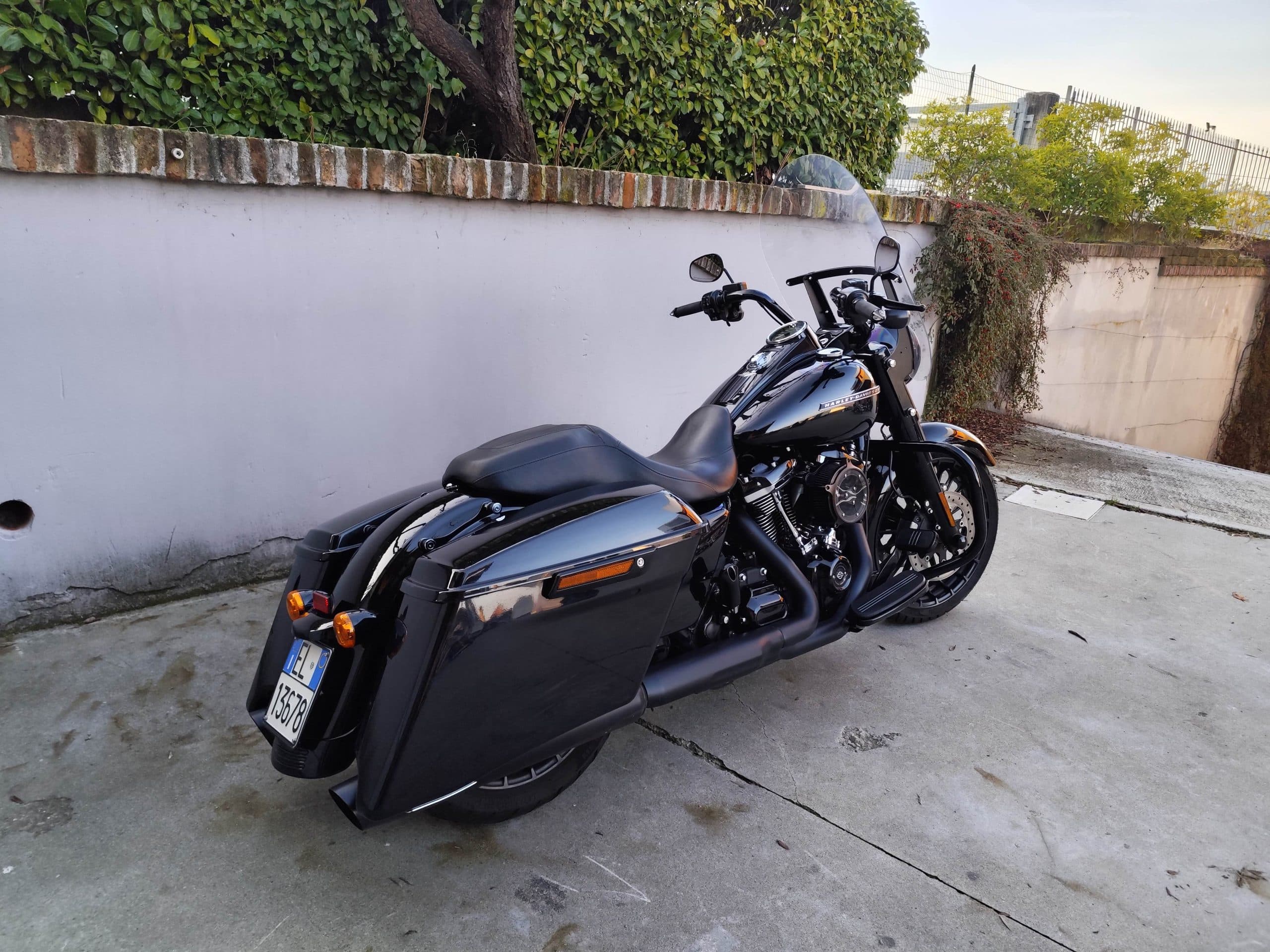 Harley-Davidson nera, modello touring con borse laterali e parabrezza, parcheggiata su pavimentazione in cemento accanto a un muro con siepe.