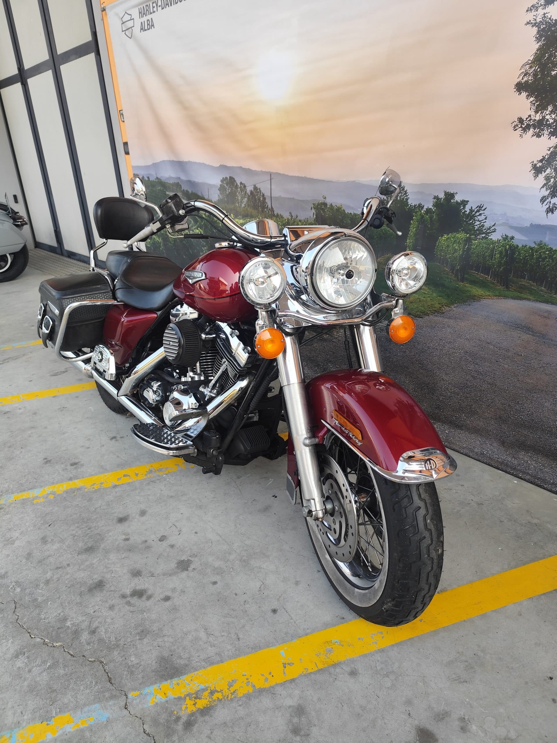 Harley-Davidson Road King rossa lucida con dettagli cromati, parcheggiata su pavimentazione in cemento. Sfondo banner promozionale Harley-Davidson Alba.