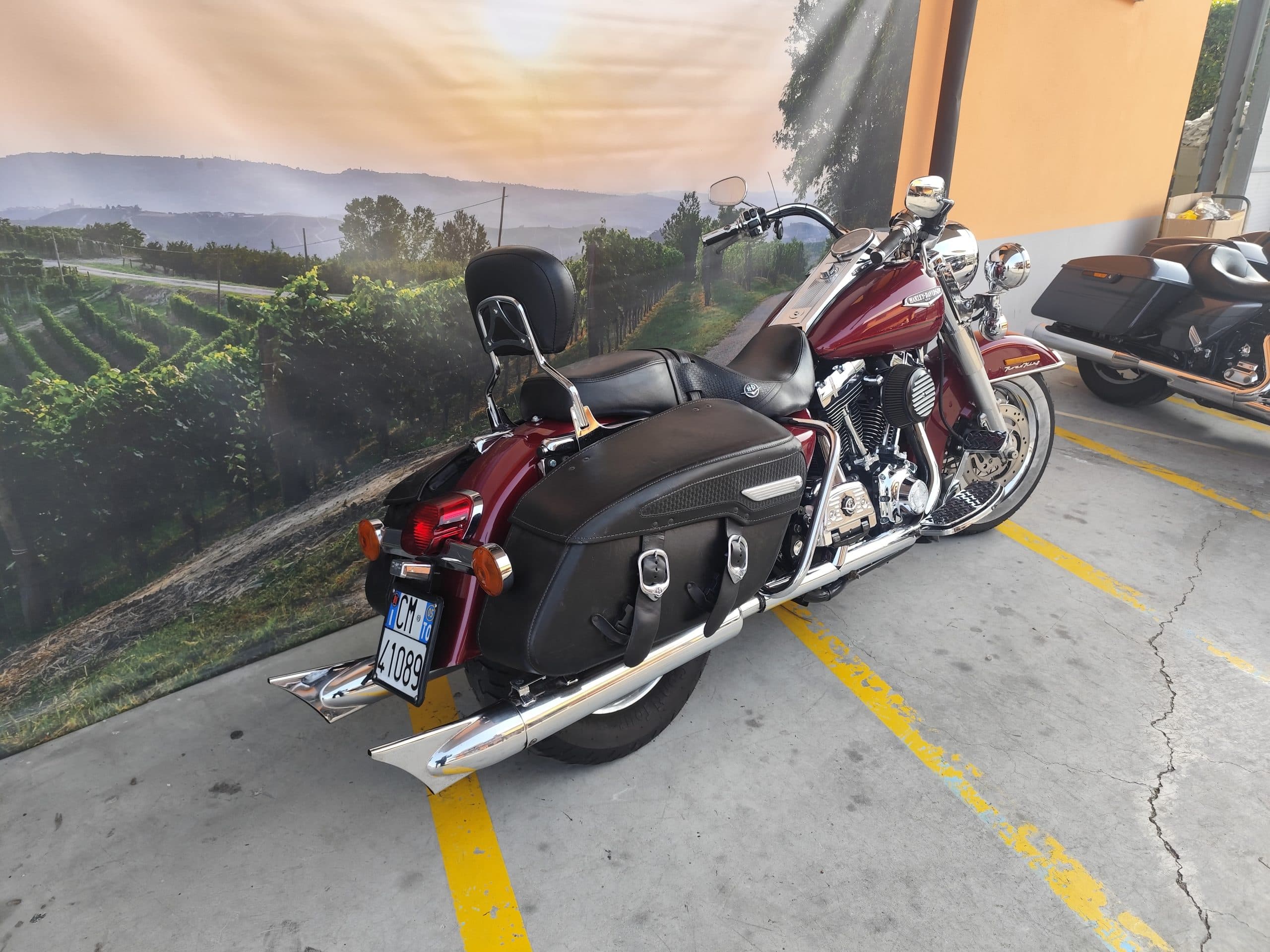Harley-Davidson Road King rossa parcheggiata su asfalto, con borse in pelle e dettagli cromati, sullo sfondo un murale di vigneti e colline al tramonto.