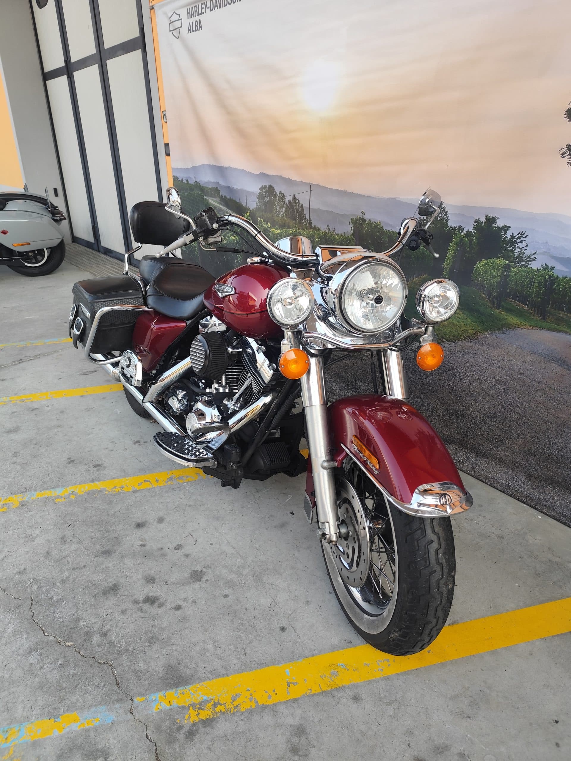 Moto Harley-Davidson rossa con dettagli cromati, parcheggiata su pavimentazione in cemento. Sullo sfondo, banner con paesaggio e logo Harley-Davidson Alba.
