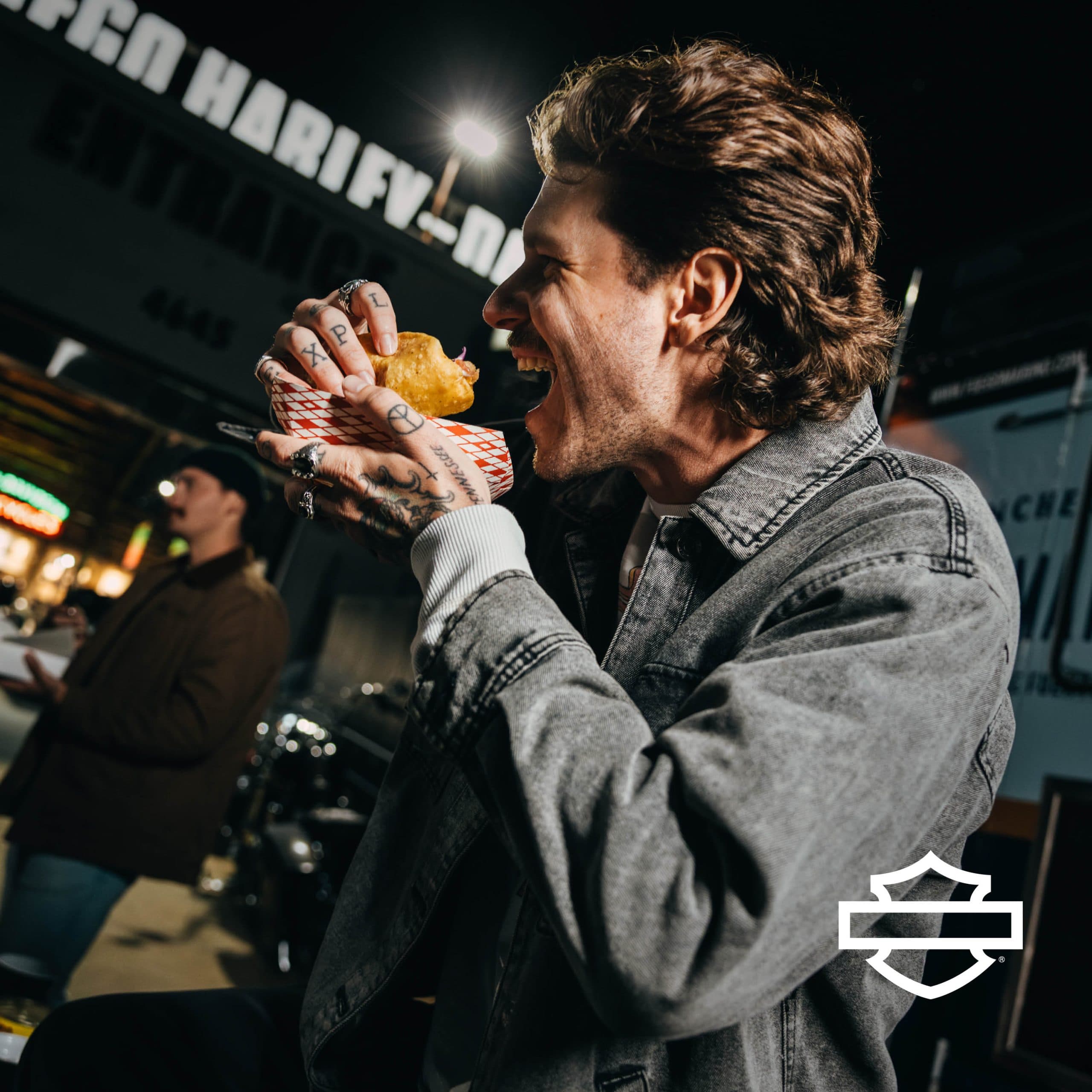 Uomo con giacca in denim e tatuaggi, intento a mordere un taco, con insegna Harley-Davidson sullo sfondo e logo del brand. Rappresenta lo stile di vita dinamico.