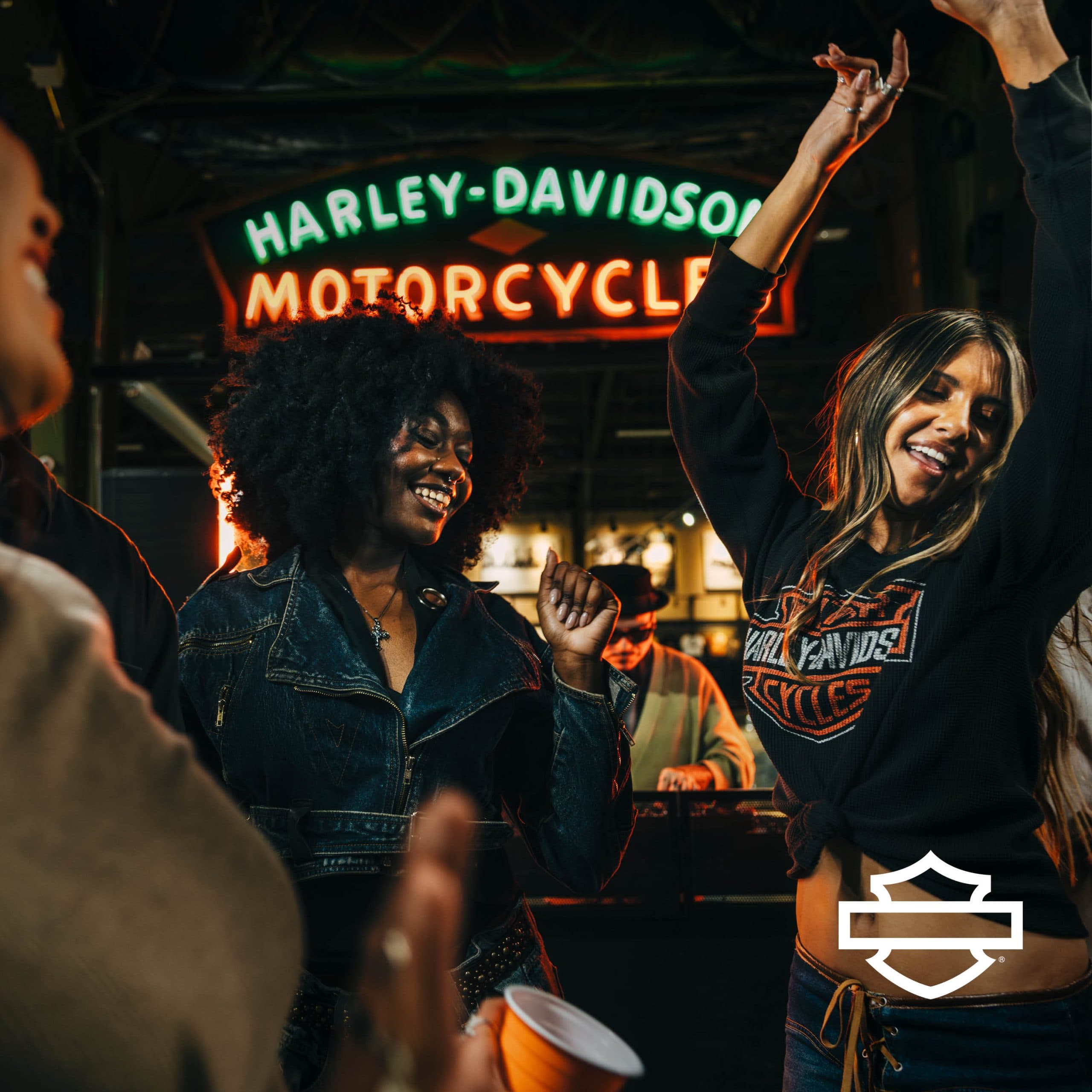 Giovani festeggiano in un locale con insegna neon Harley-Davidson Motorcycle. Abbigliamento brandizzato Harley-Davidson, espressione dello stile di vita.