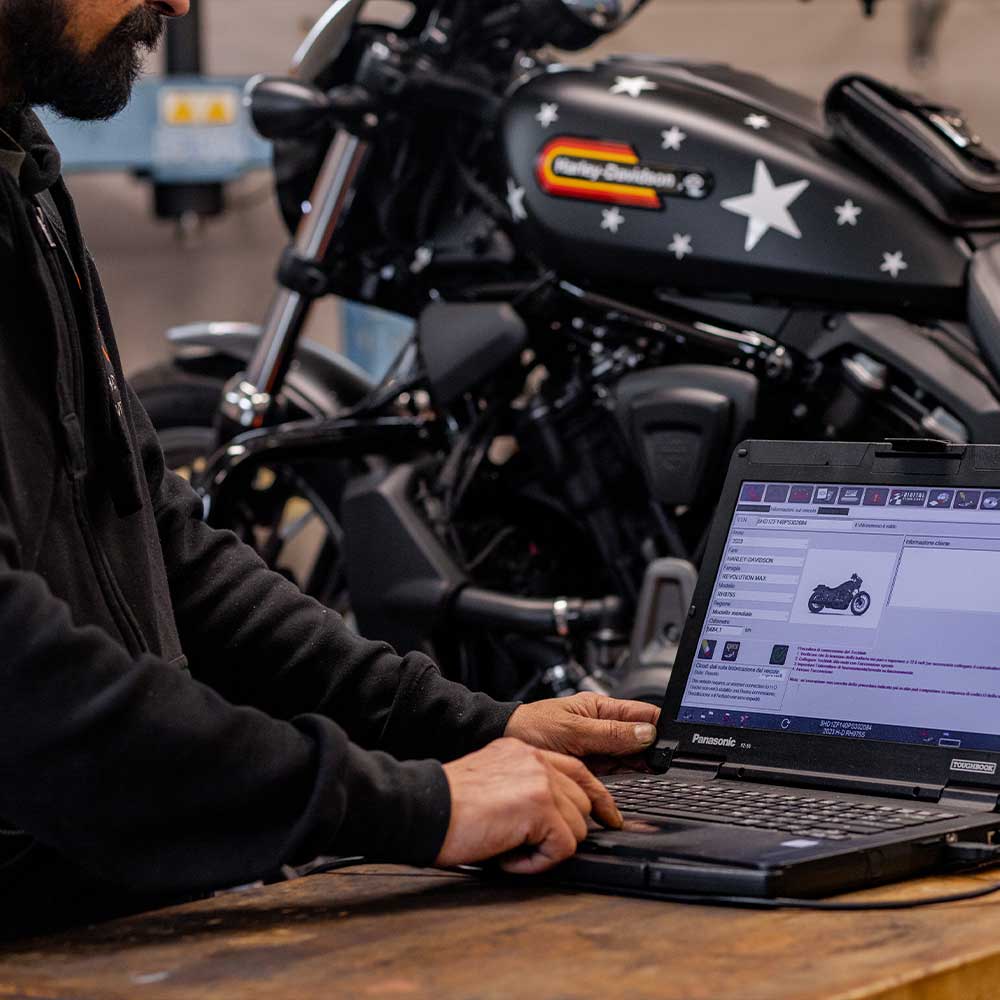 Tecnico Harley-Davidson esegue diagnostica elettronica su moto con laptop Panasonic Toughbook, visualizzando dati motore Revolution Max.