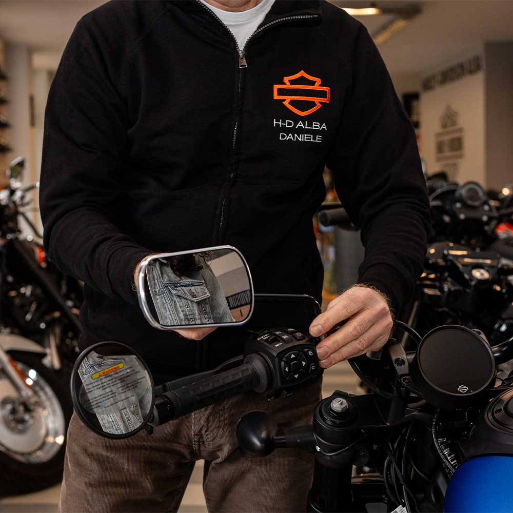 Daniele di H-D Alba, con felpa Harley-Davidson, regola i comandi di una moto. I retrovisori riflettono una giacca in denim, suggerendo un test ride.