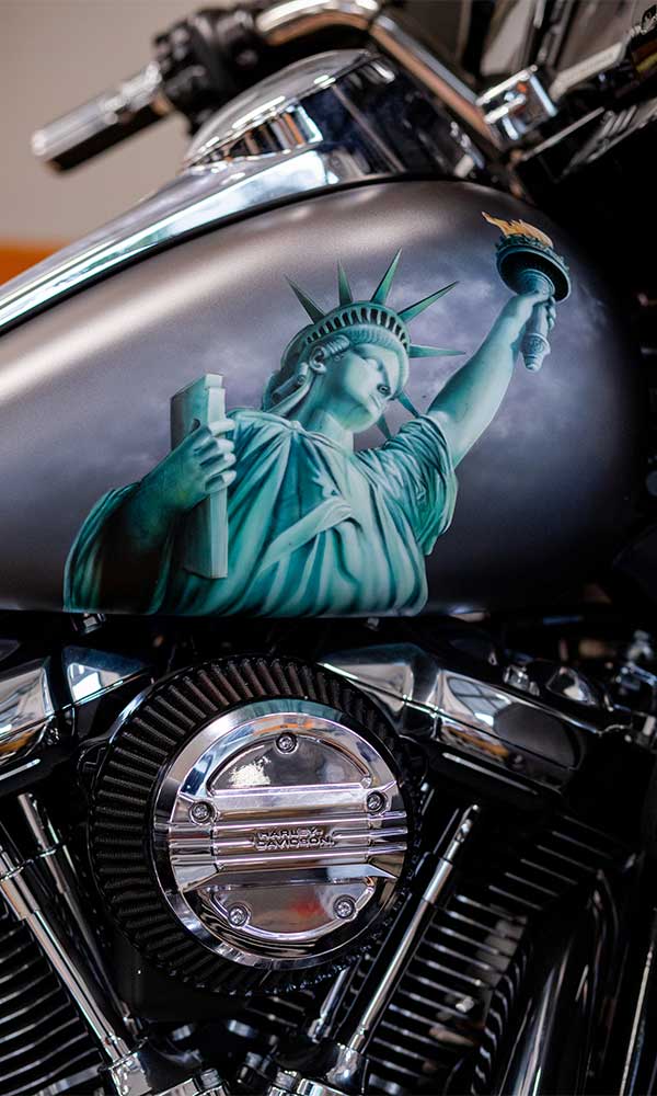 Serbatoio carburante moto Harley-Davidson personalizzato con aerografia Statua della Libertà. Dettaglio motore cromato con logo Harley-Davidson.
