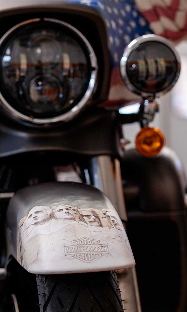 Dettaglio frontale di moto Harley-Davidson personalizzata. Faro LED, parafango anteriore con decorazione Mount Rushmore e logo, e bandiera USA.