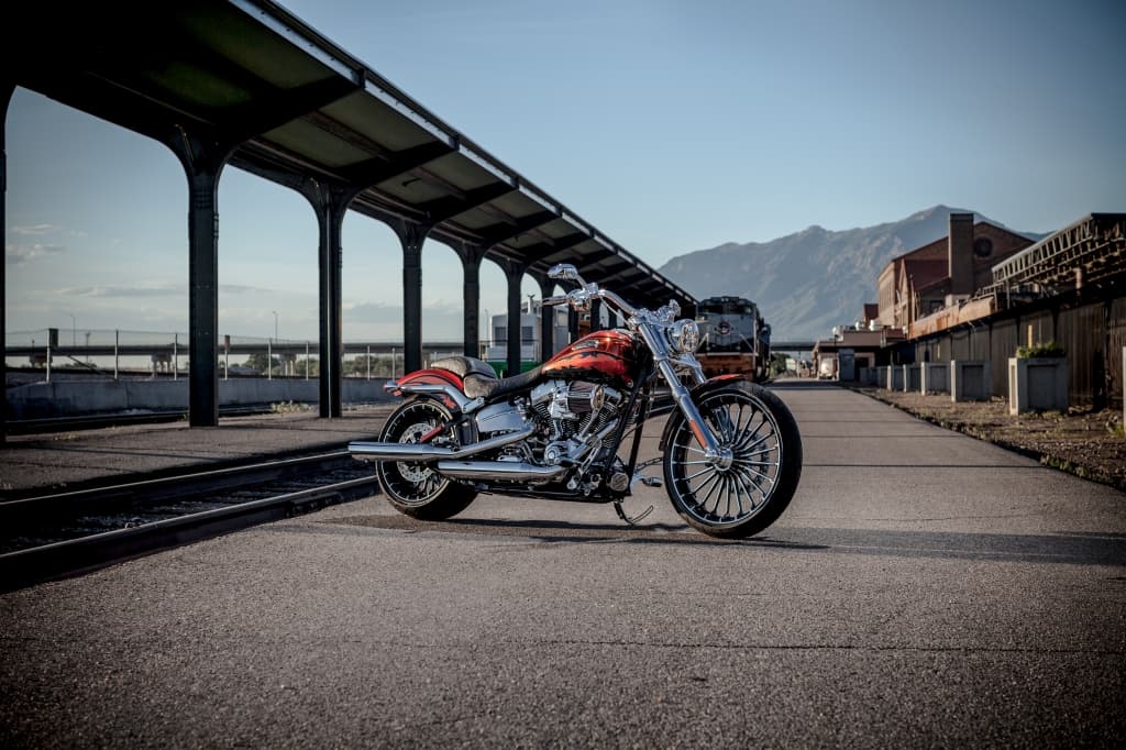 Harley-Davidson customizzata con livrea arancione fiammata, motore cromato e ruote a raggi, parcheggiata su banchina ferroviaria con treno e montagne sullo sfondo.