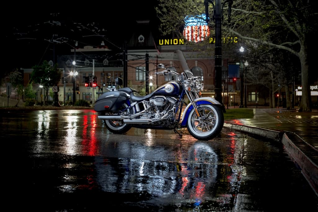Harley-Davidson blu e bianca, modello cruiser, parcheggiata su strada bagnata di notte, con riflessi di luci urbane. Dettagli cromati e pneumatici whitewall.