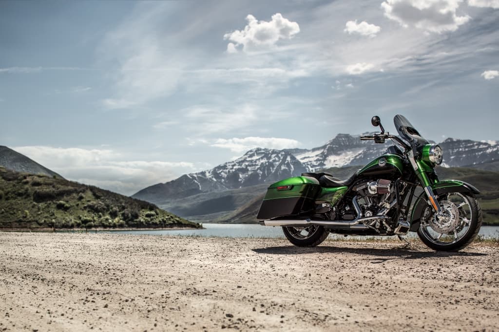 Harley-Davidson touring verde metallizzato parcheggiata su sterrato, con lago e montagne innevate sullo sfondo. Ideale per test ride avventurosi.