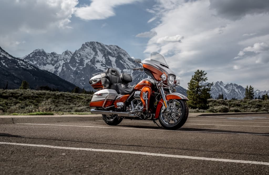Harley-Davidson touring Electra Glide arancione e bianca parcheggiata, con maestose montagne innevate e cielo nuvoloso sullo sfondo, ideale per viaggi.