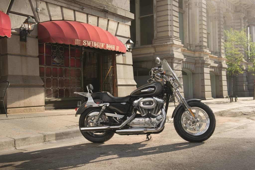 Harley-Davidson con motore V-twin, finitura scura, parabrezza e dettagli cromati, parcheggiata su strada urbana. Ideale per test ride.