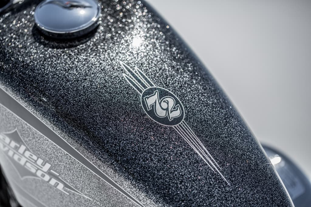 Primo piano del serbatoio carburante di una Harley-Davidson modello 72, con verniciatura metal flake grigio scuro e logo "72" in evidenza.