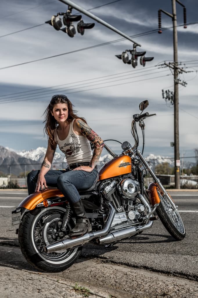 Motociclista con abbigliamento Harley-Davidson e tatuaggi, seduta su una moto Harley-Davidson Sportster arancione. Sfondo montano. Perfetta per un test ride.