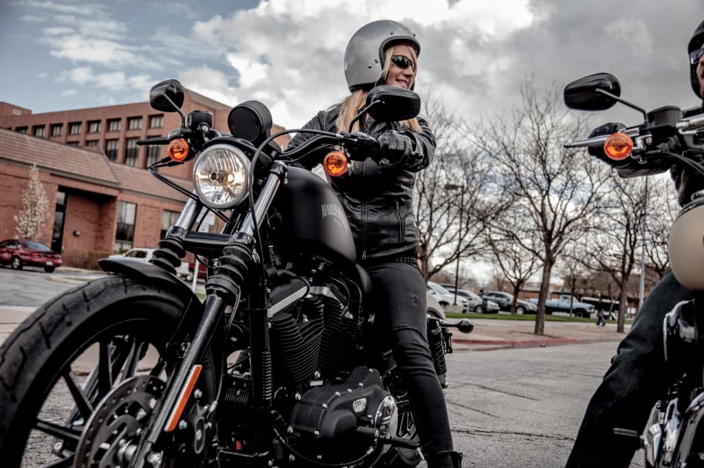 Donna con casco e abbigliamento tecnico su moto Harley-Davidson nera, motore in evidenza, pronta per un test ride. Altro motociclista.