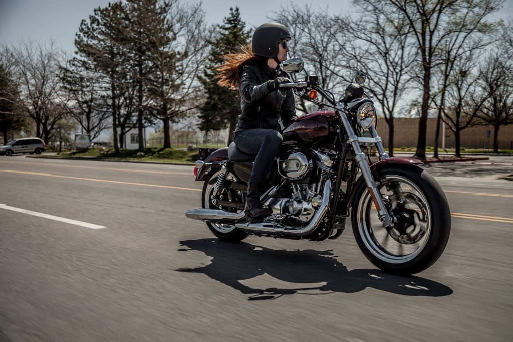 Donna con casco e giacca in pelle nera alla guida di una Harley-Davidson bordeaux su strada asfaltata, con alberi spogli sullo sfondo.