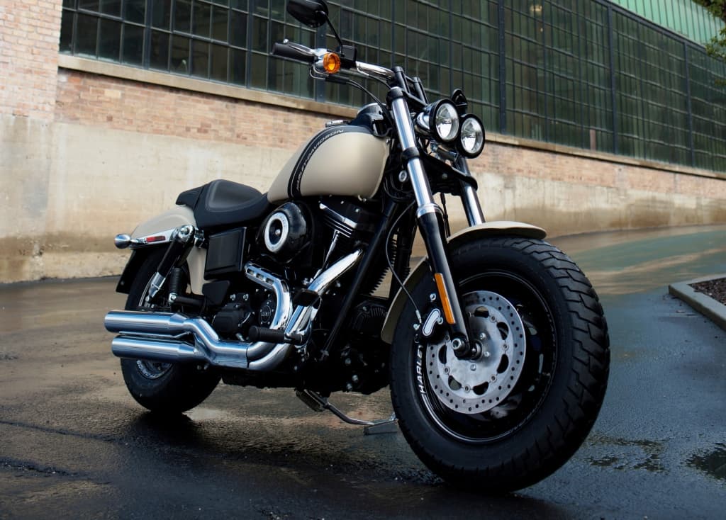 Harley-Davidson Fat Bob con livrea beige e nera, pneumatici larghi e doppi fari, parcheggiata su asfalto bagnato. Ideale per un test ride.