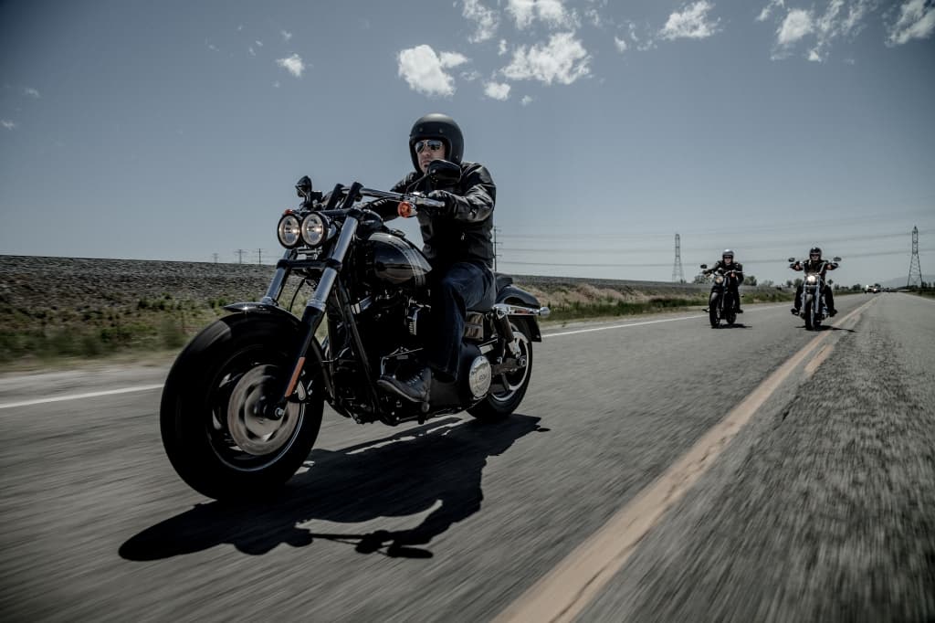 Tre motociclisti su Harley-Davidson in test ride su strada aperta. Il pilota in primo piano indossa casco e abbigliamento tecnico da moto.