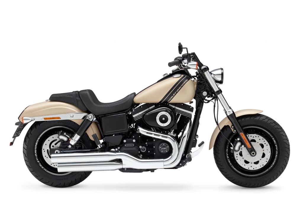 Harley-Davidson Fat Bob color sabbia con motore nero, scarichi cromati, pneumatici larghi e doppio freno a disco anteriore, su sfondo bianco.