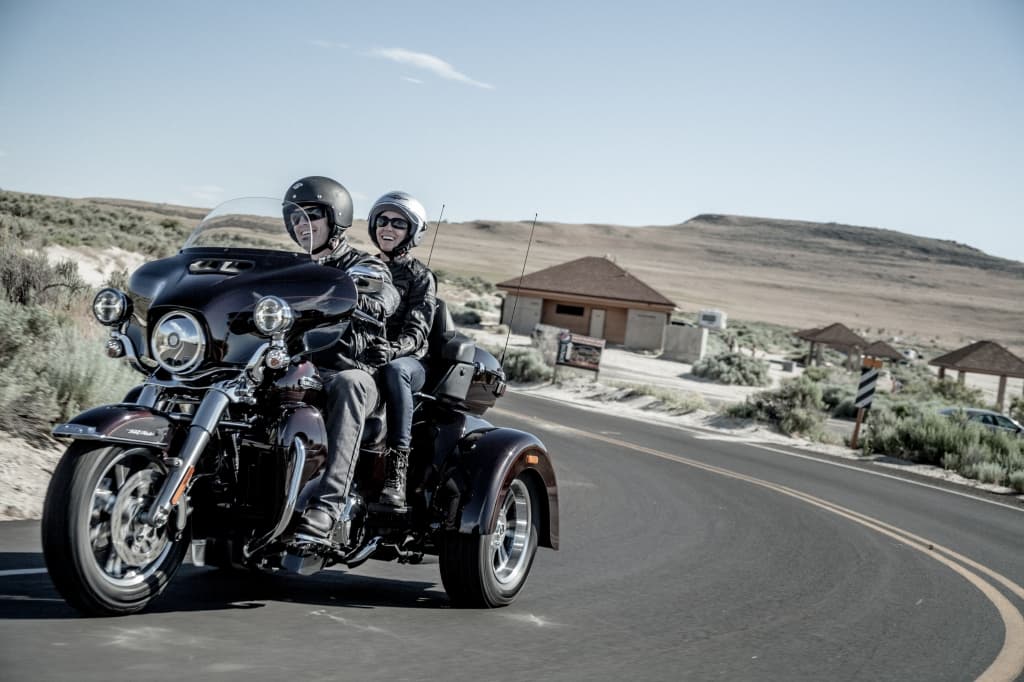 Coppia in abbigliamento Harley-Davidson su Trike, sorridente durante un test ride o viaggio su strada panoramica.
