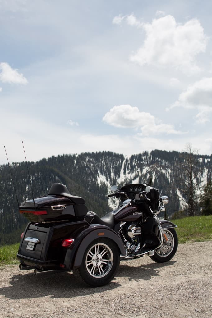 Harley-Davidson Tri Glide Ultra bordeaux parcheggiato su strada sterrata, con montagne innevate e cielo nuvoloso sullo sfondo.