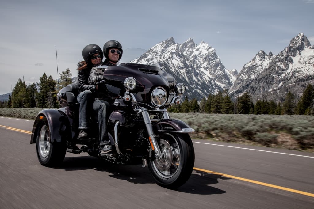 Coppia sorridente su trike Harley-Davidson scuro, con abbigliamento da moto, in corsa su strada panoramica con maestose montagne innevate.