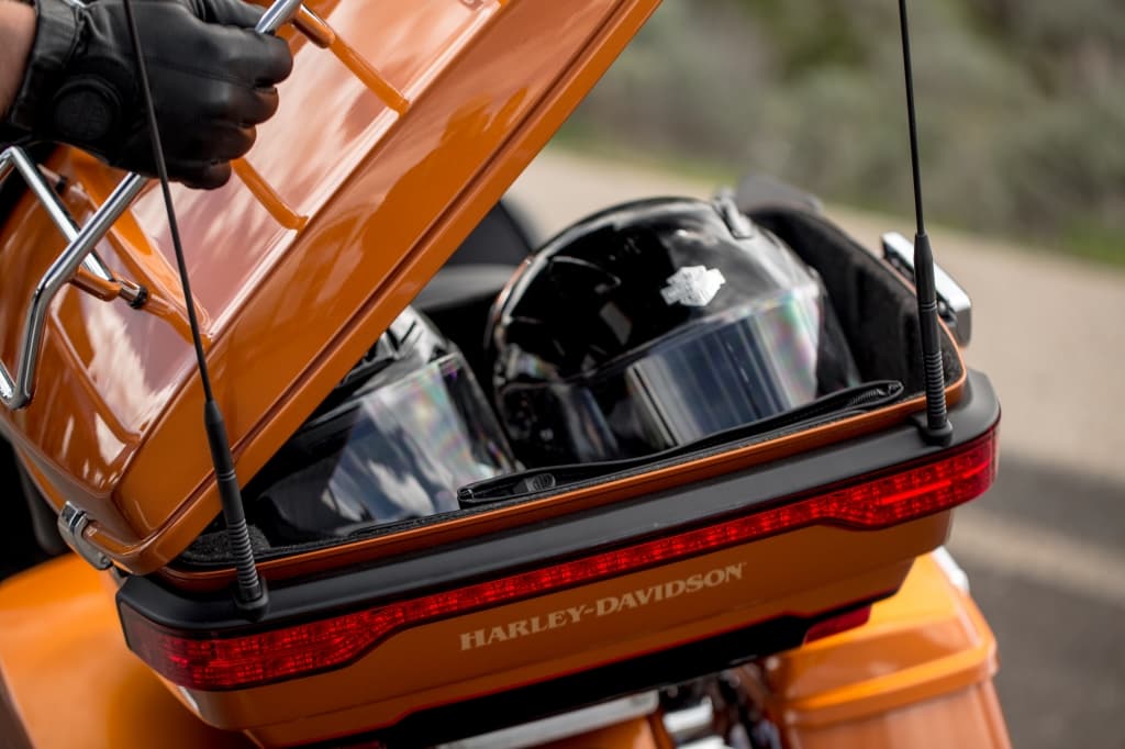 Bauletto posteriore arancione di moto Harley-Davidson aperto, contenente due caschi neri. Una mano guantata tiene il coperchio. Dettaglio del logo Harley-Davidson.