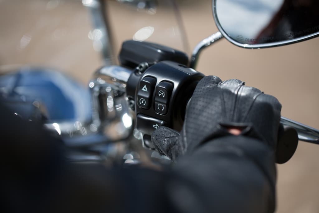 Mano guantata in pelle nera sulla manopola destra di una Harley-Davidson, con comandi per frecce, hazard e cruise control. Test ride.