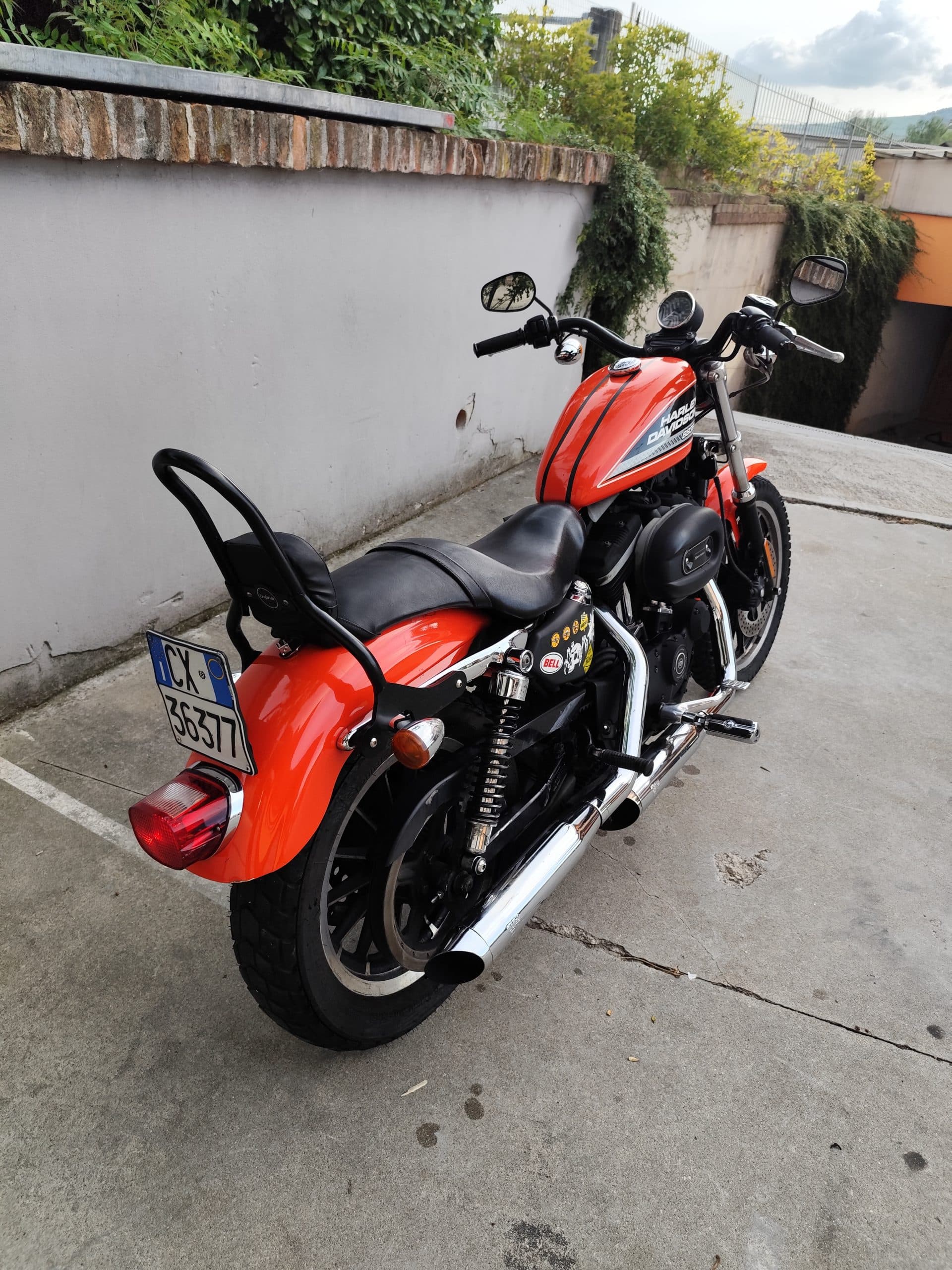 Harley-Davidson Sportster arancione parcheggiata, vista posteriore-laterale destra, con targa italiana, sissy bar e scarichi cromati.