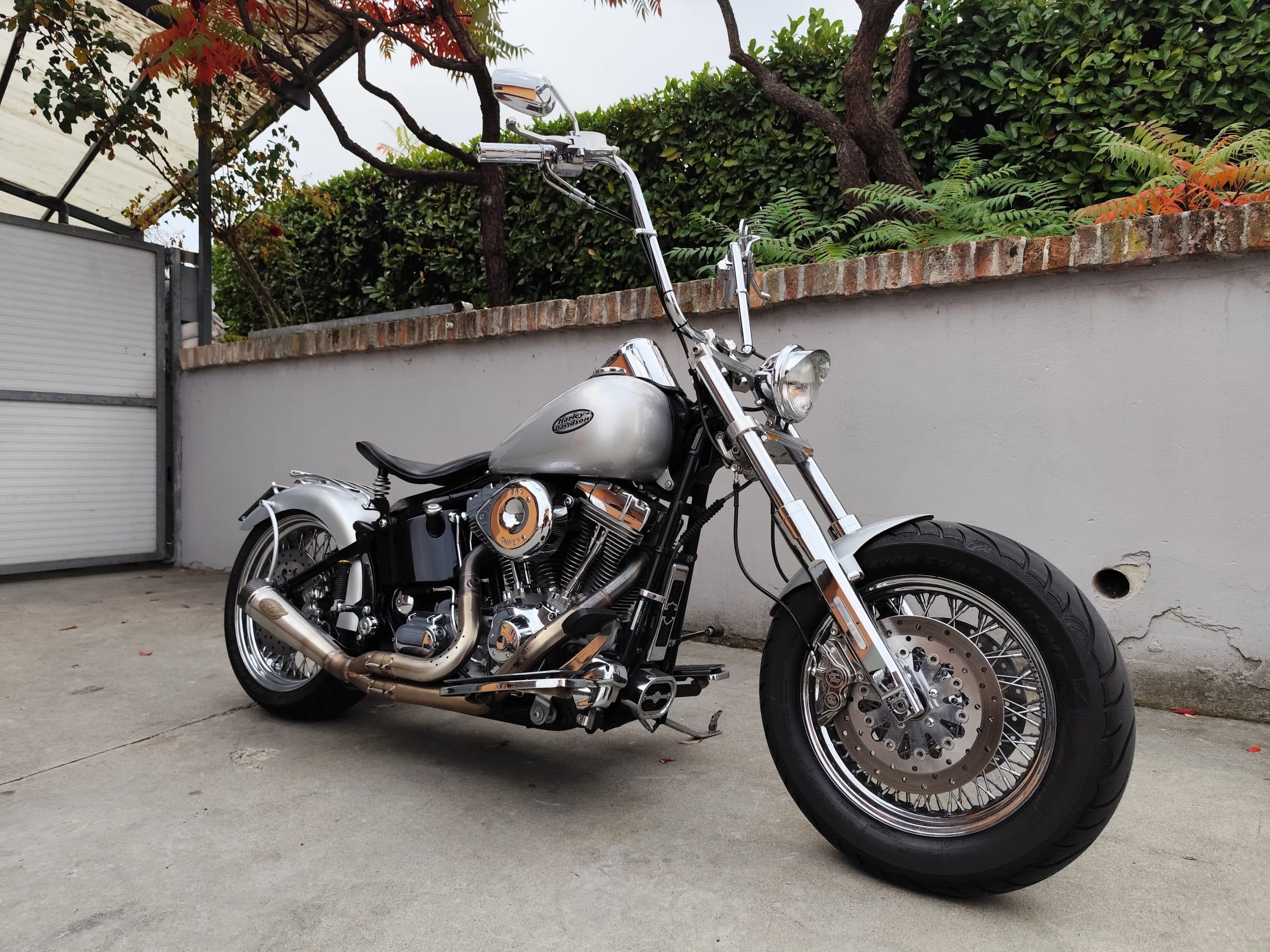 Harley-Davidson customizzata argento con manubrio ape hanger, motore V-twin cromato, scarico after-market e ruote a raggi, su pavimentazione.