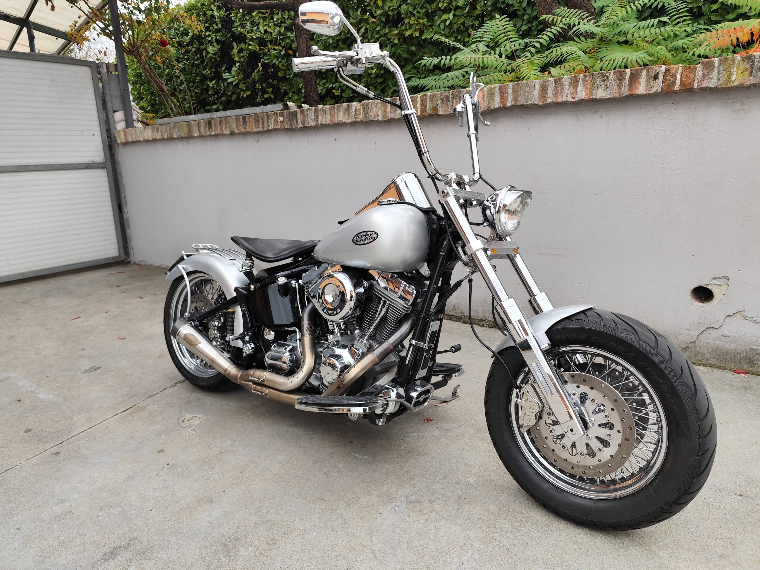 Harley-Davidson customizzata argento con manubrio ape hanger, motore cromato S&S, scarico Cobra e ruote a raggi, parcheggiata.