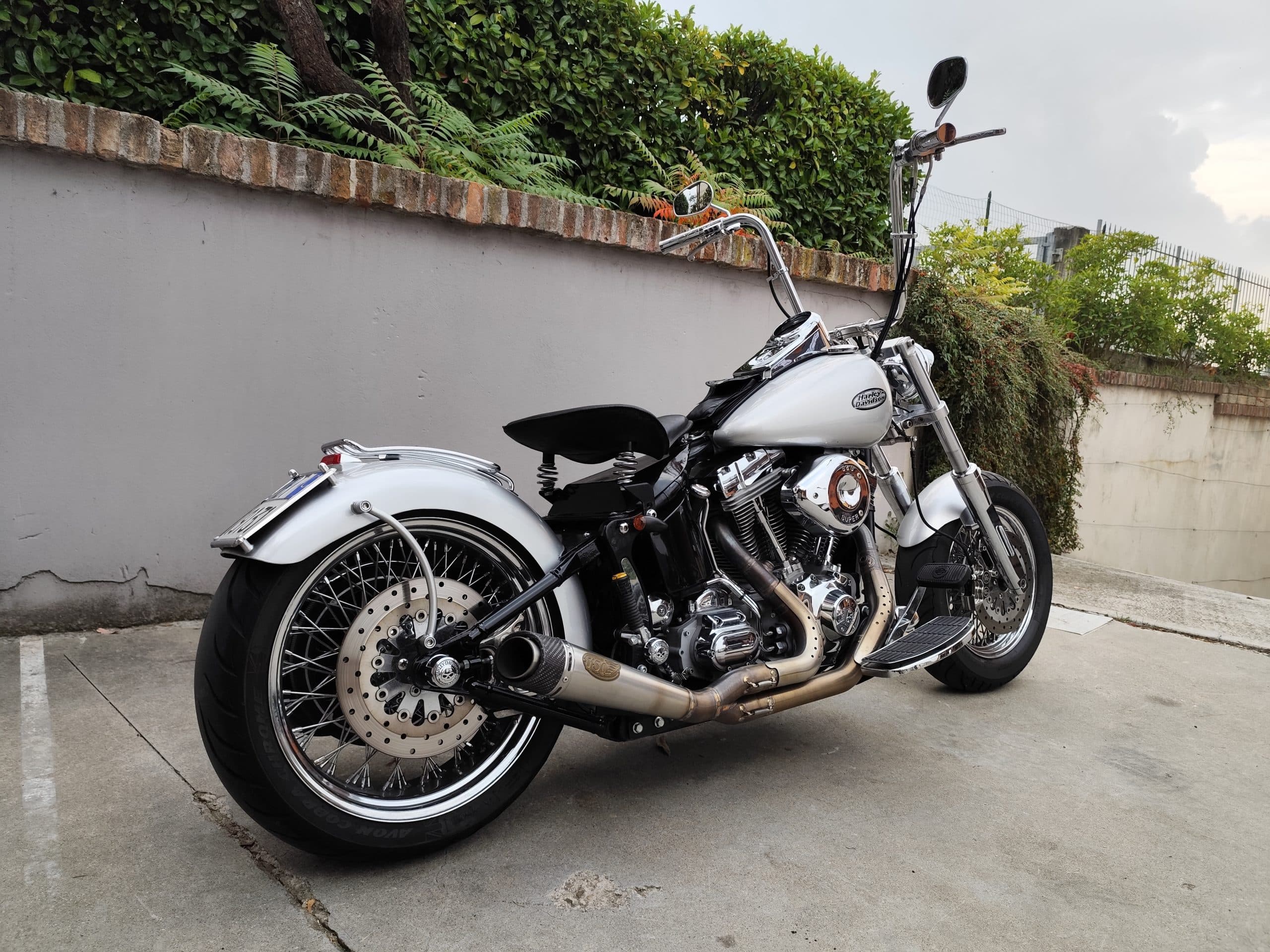 Motocicletta Harley-Davidson customizzata, colore argento metallizzato, con scarico aftermarket e sella monoposto a molle, parcheggiata su cemento.