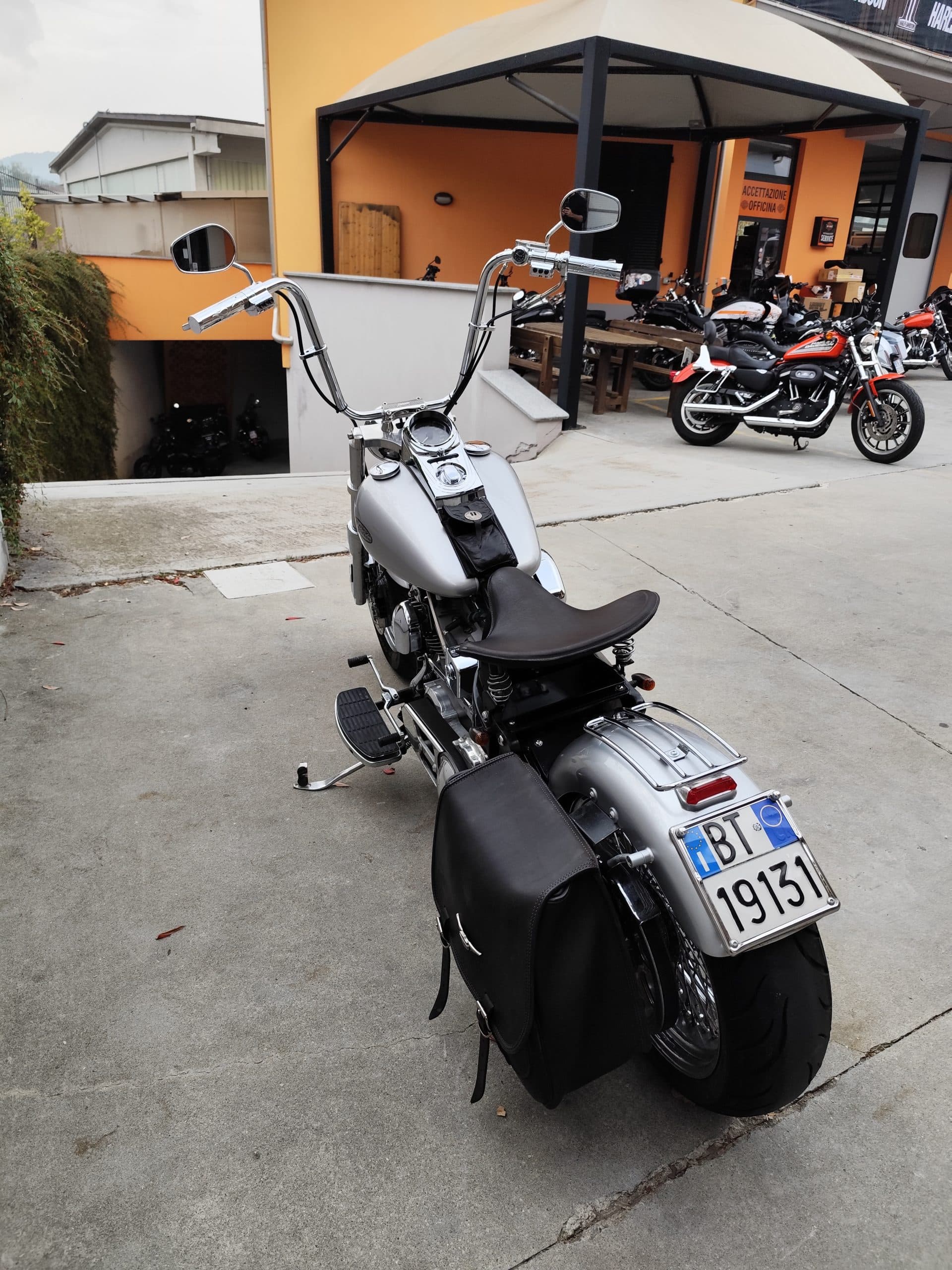 Harley-Davidson custom cromata con manubrio alto e borse laterali, parcheggiata davanti alla concessionaria con officina e altri modelli.