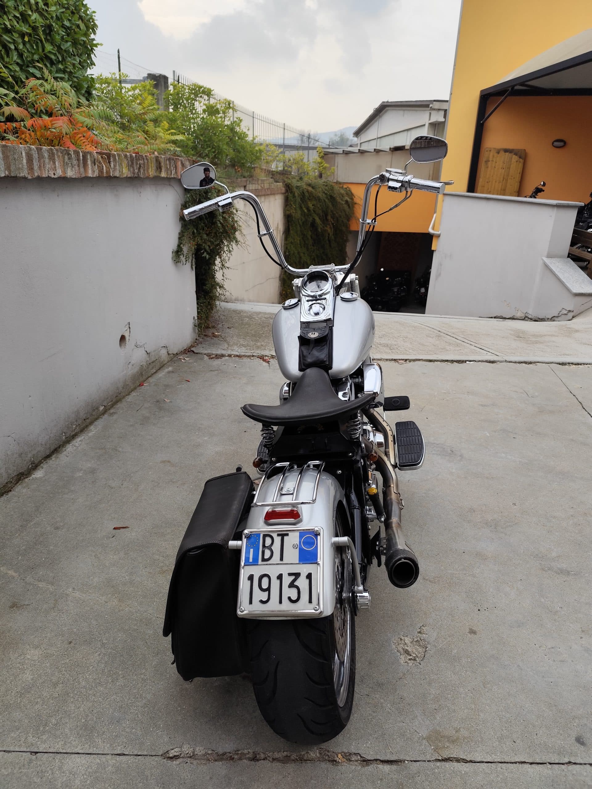 Harley-Davidson customizzata argento con manubrio ape hanger, sella monoposto nera e targa italiana BT 19131, parcheggiata all'aperto.