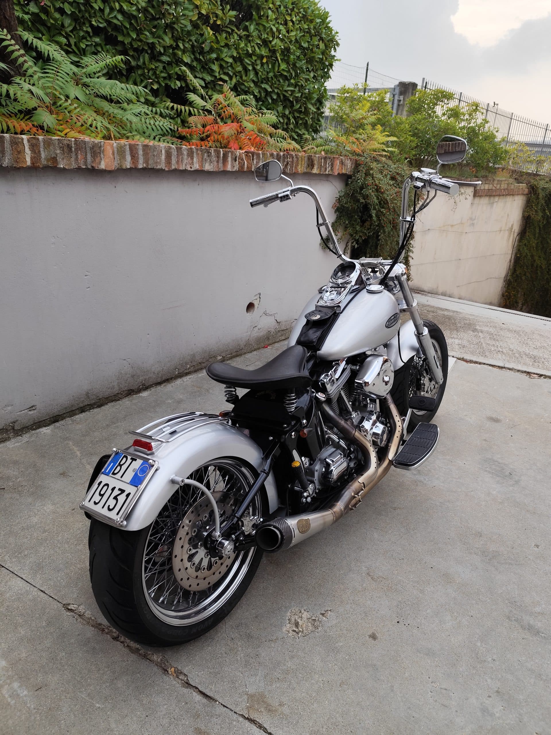 Harley-Davidson customizzata argento con manubrio ape-hanger, sella monoposto a molle, scarico personalizzato e ruote a raggi, parcheggiata su cemento.