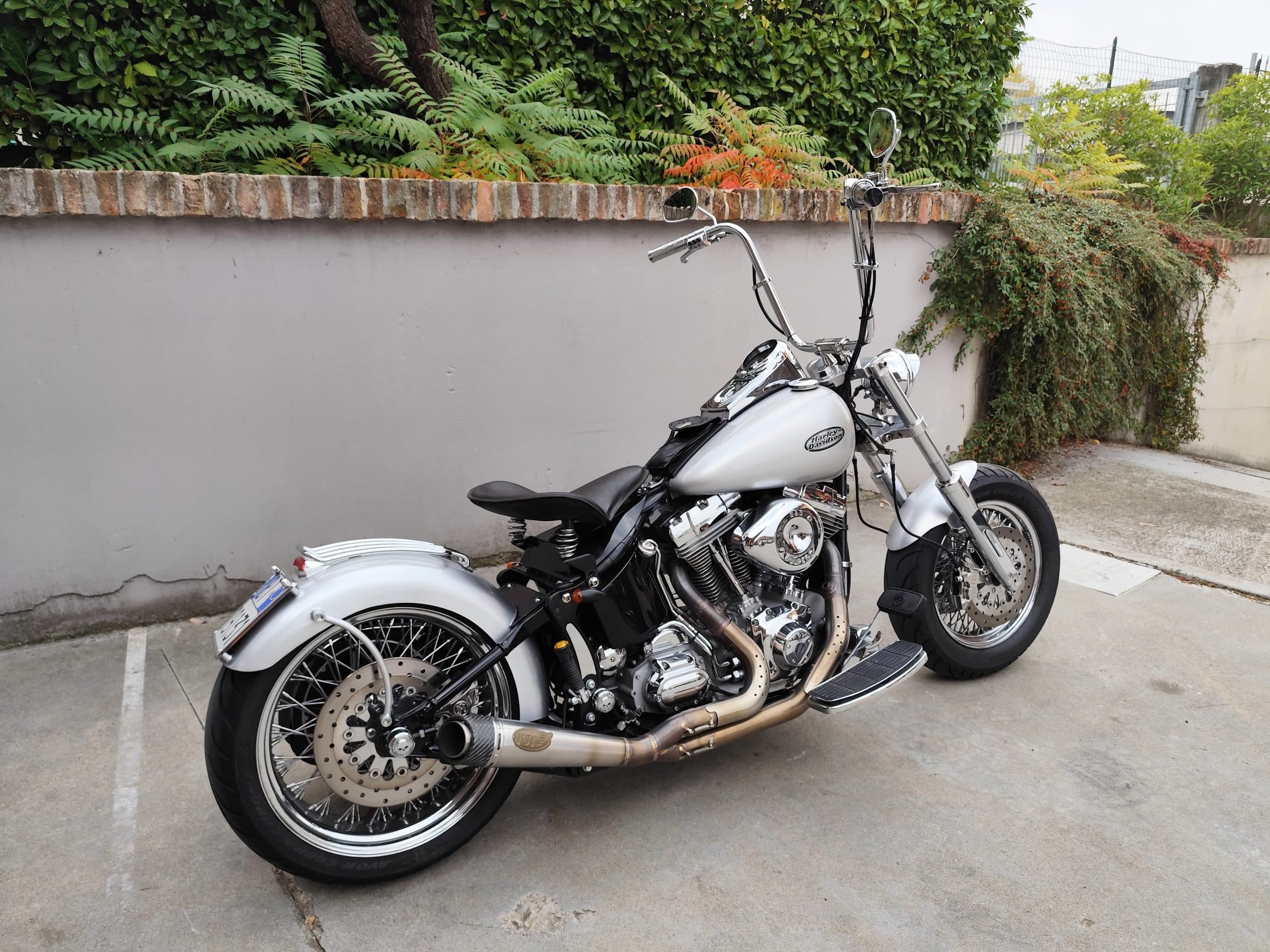 Harley-Davidson customizzata argento con manubrio ape hanger, motore cromato, scarico Vance & Hines e ruote a raggi, parcheggiata all'aperto.