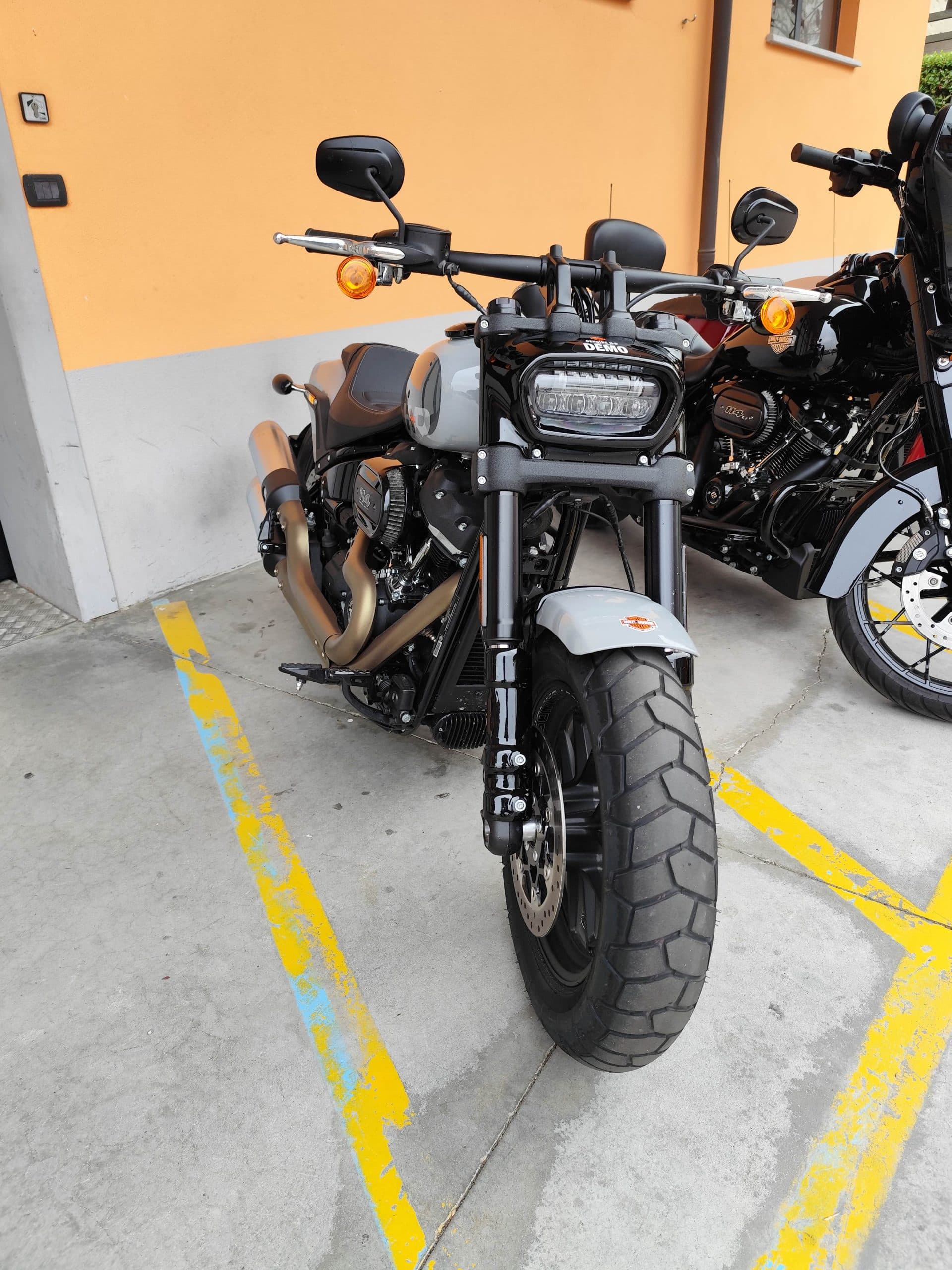 Harley-Davidson grigia, modello test ride con scritta DEMO sul faro, parcheggiata su asfalto con segnaletica gialla, affiancata da moto nera.