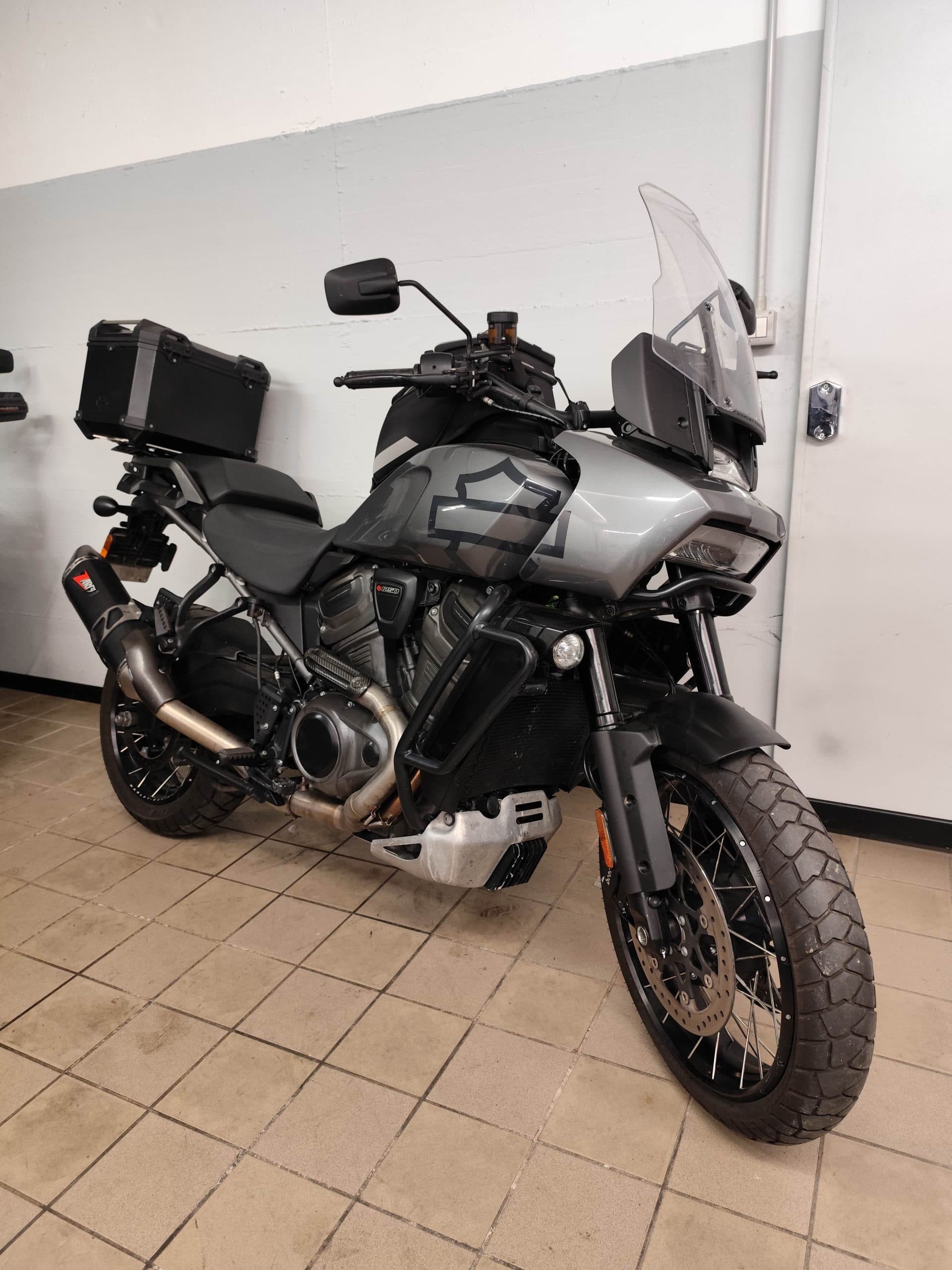 Harley-Davidson Pan America 1250 Special grigia con bauletto, scarico Akrapovic e pneumatici tassellati, parcheggiata in concessionaria.