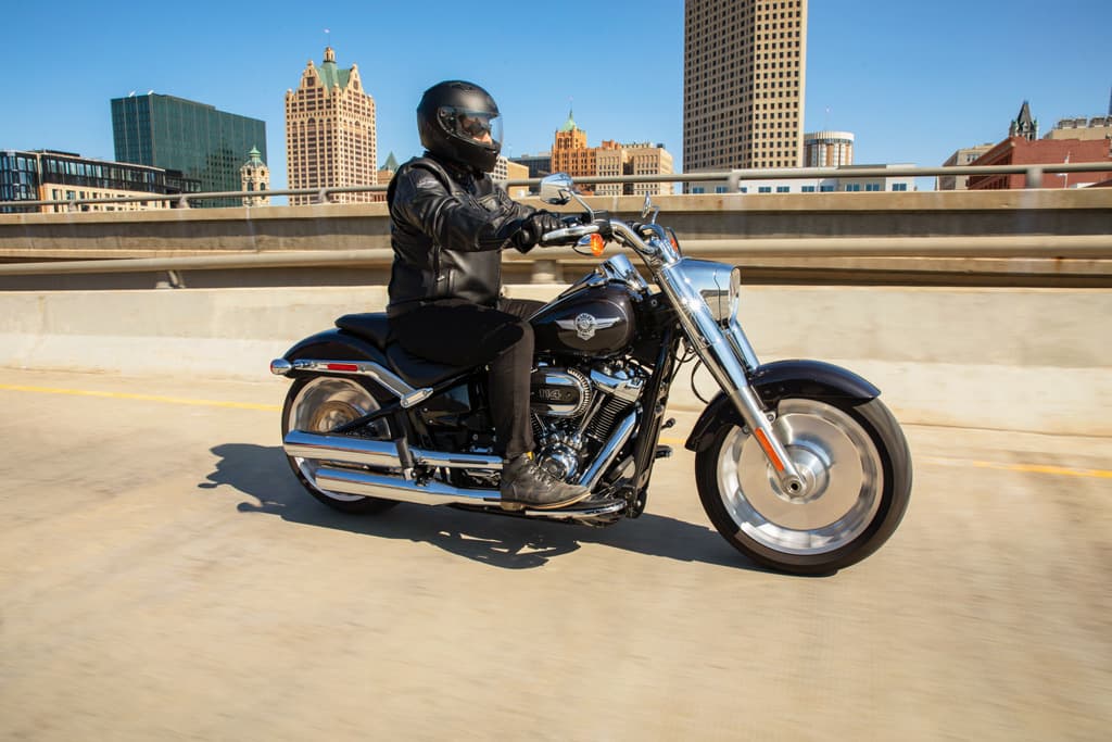 Pilota in abbigliamento tecnico su moto Harley-Davidson nera con motore 114, in test ride su strada urbana con skyline.