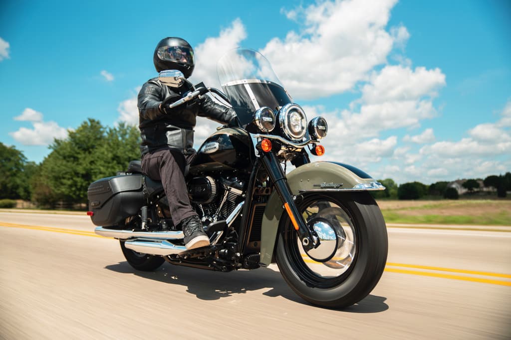 Motociclista con casco integrale e giacca in pelle nera in sella a una Harley-Davidson Heritage Classic bicolore su strada. Test ride.