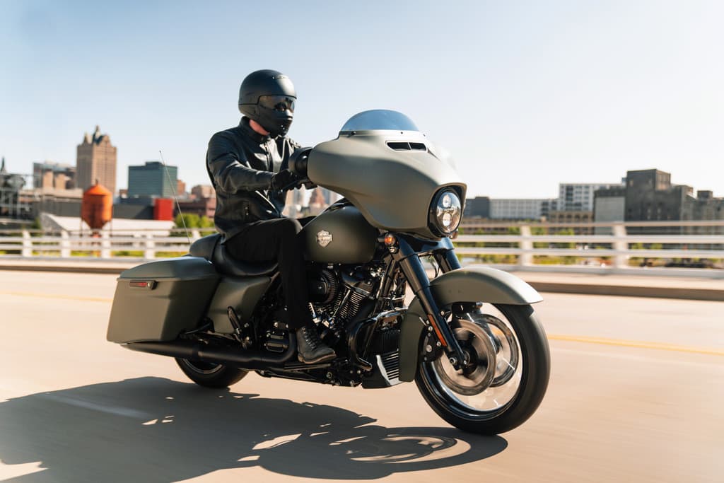 Motociclista con abbigliamento tecnico su Harley-Davidson Street Glide verde opaco in test ride su strada panoramica con skyline urbano.
