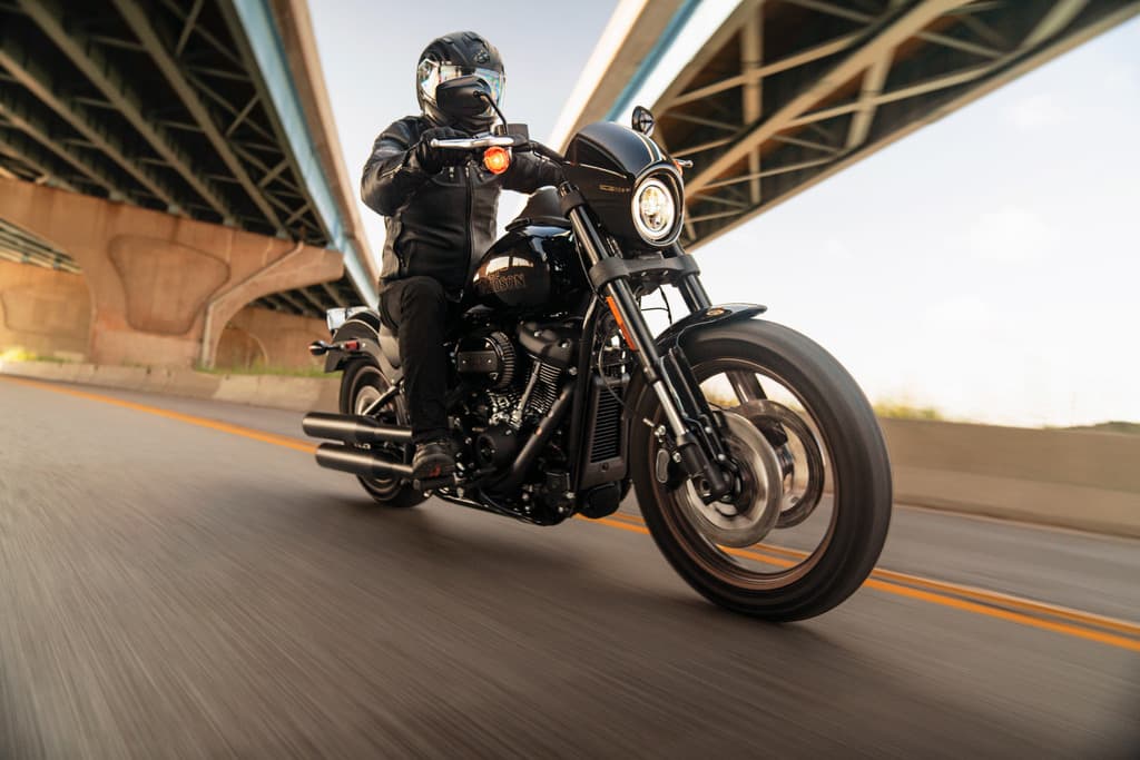 Motociclista con abbigliamento tecnico e casco guida una Harley-Davidson nera su strada. L'effetto mosso suggerisce un test ride dinamico.