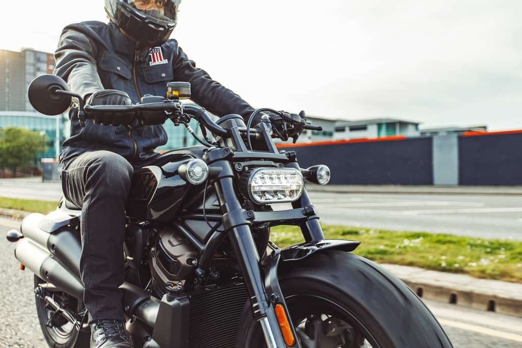 Motociclista con casco e giacca in pelle con patch USA su moto Harley-Davidson nera. Dettagli di abbigliamento tecnico e motore in evidenza durante un test ride.