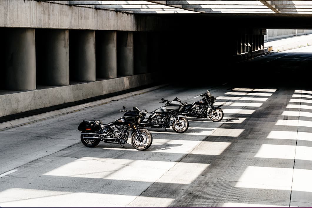 Tre motociclette Harley-Davidson scure, allineate sotto un cavalcavia in cemento, con giochi di luce e ombra che ne esaltano il design.