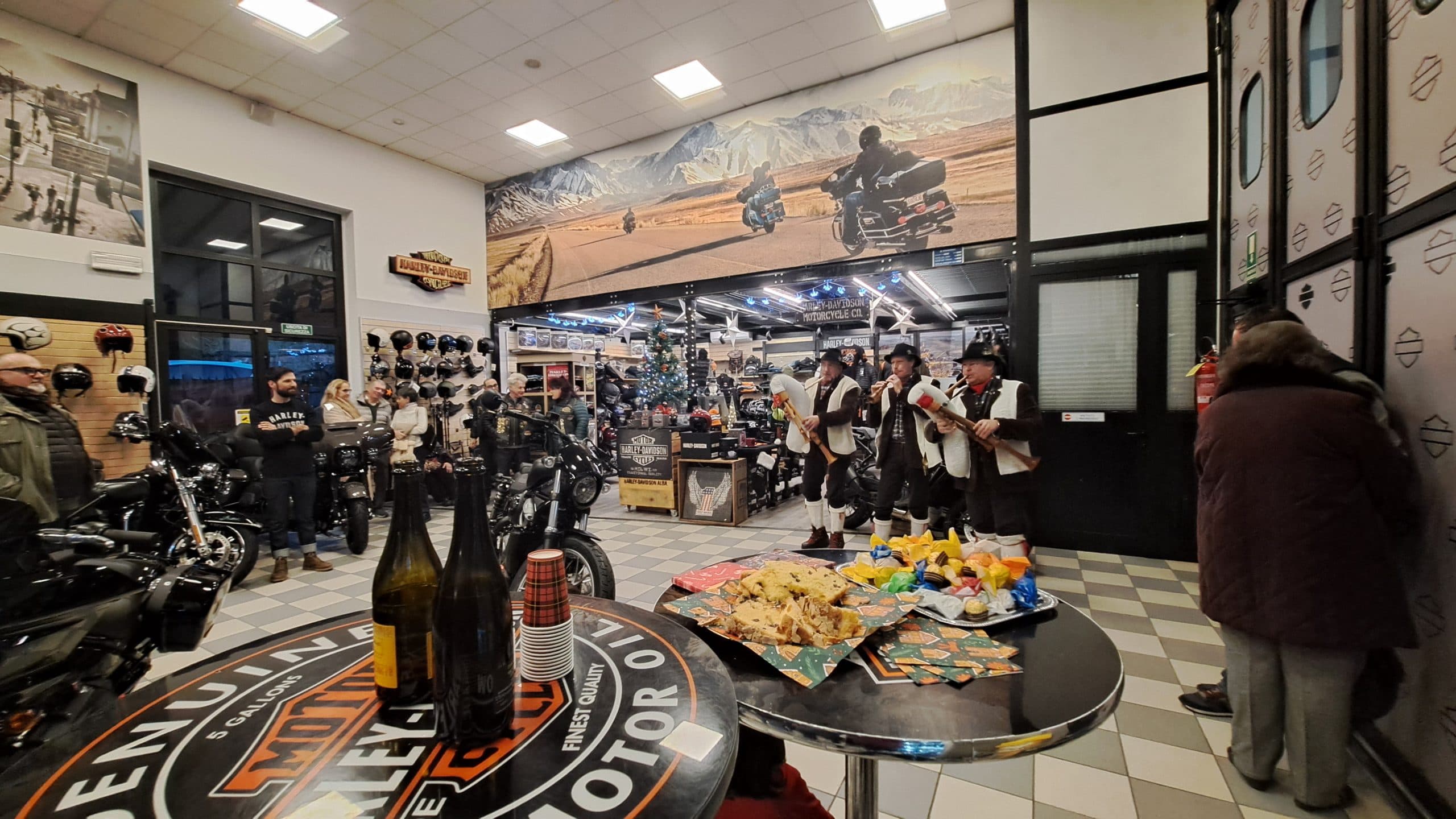 Evento festivo presso la concessionaria Harley-Davidson Alba con esposizione di moto, pubblico, musicisti tradizionali e buffet.