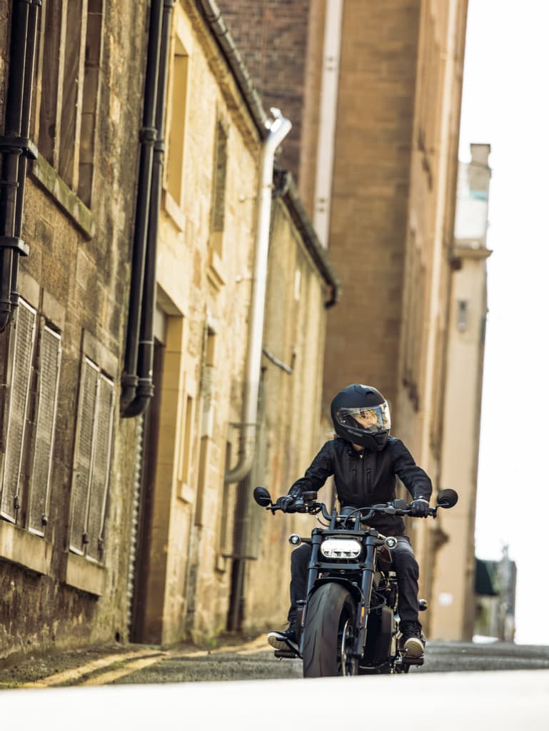 Motociclista con casco integrale e abbigliamento tecnico nero su una Harley-Davidson moderna, in una via urbana stretta. Ideale per test ride.