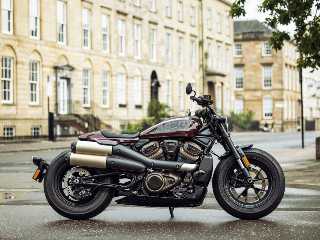 Moto Harley-Davidson Sportster S color bordeaux su strada bagnata, con architettura classica. Scopri i motori e prenota un test ride.